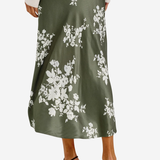 A woman wearing a midi-length olive green satin skirt with a white floral print, styled with a cream long-sleeve top and black strappy high-heeled sandals against a light gray background.