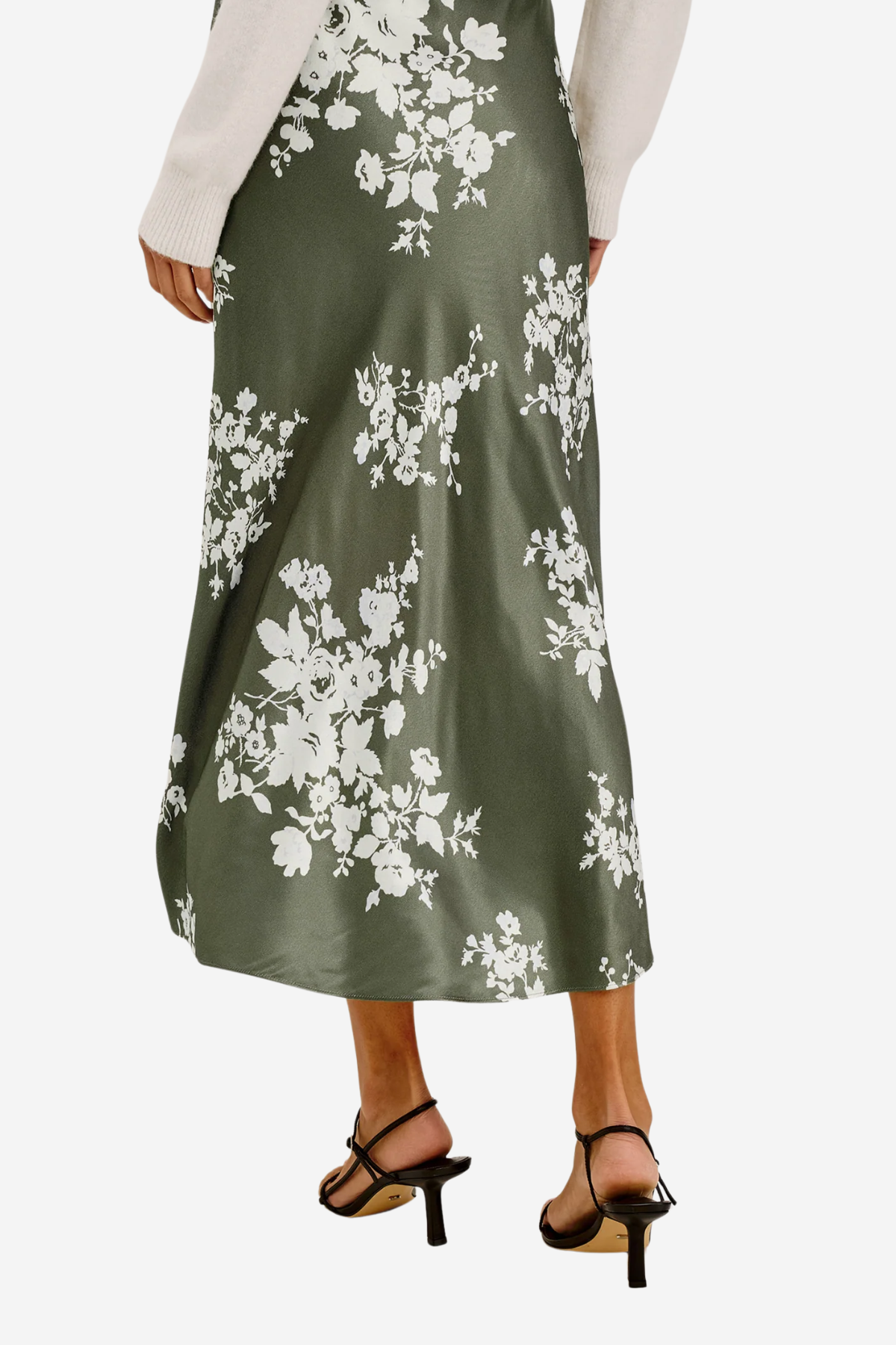 A woman wearing a midi-length olive green satin skirt with a white floral print, styled with a cream long-sleeve top and black strappy high-heeled sandals against a light gray background.
