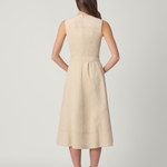 A woman viewed from the back wearing a sleeveless, knee-length beige dress with a gathered waist and brown ankle boots, standing against a light gray studio background.