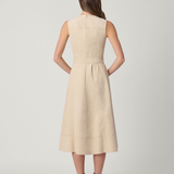 A woman viewed from the back wearing a sleeveless, knee-length beige dress with a gathered waist and brown ankle boots, standing against a light gray studio background.