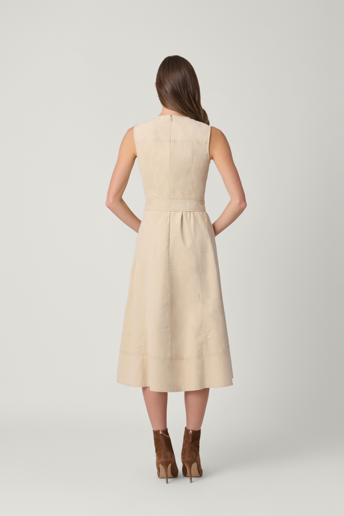 A woman viewed from the back wearing a sleeveless, knee-length beige dress with a gathered waist and brown ankle boots, standing against a light gray studio background.