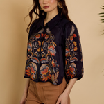 A woman modeling a dark navy blue satin blouse with intricate multicolor floral and butterfly embroidery, paired with brown pants, set against a warm beige background.