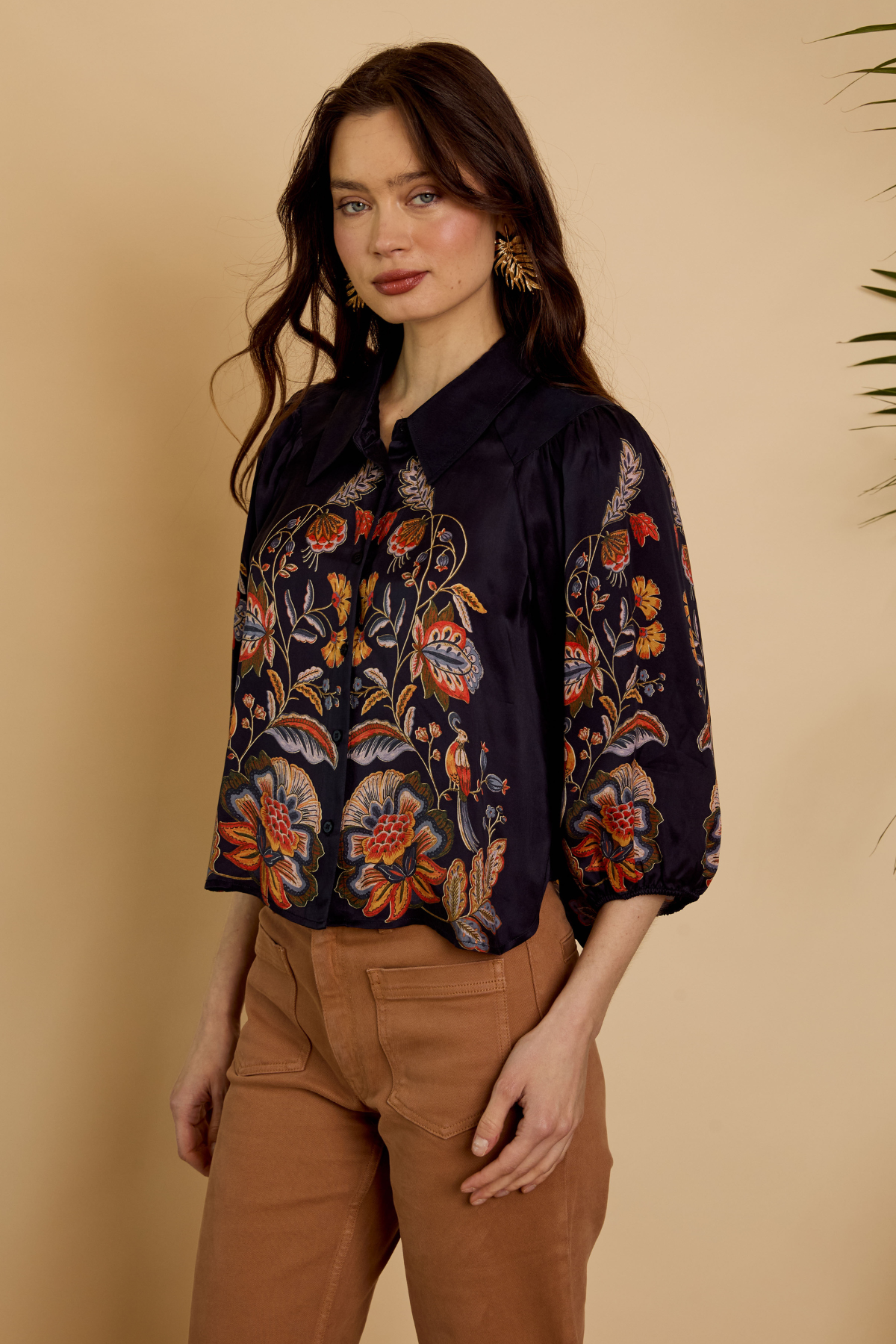 A woman modeling a dark navy blue satin blouse with intricate multicolor floral and butterfly embroidery, paired with brown pants, set against a warm beige background.