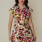 A woman modeling a short-sleeve collared dress with a bold retro leaf pattern in red, brown, olive green, and cream on a light background, set against a plain beige studio backdrop.