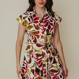 A woman modeling a short-sleeve collared dress with a bold retro leaf pattern in red, brown, olive green, and cream on a light background, set against a plain beige studio backdrop.