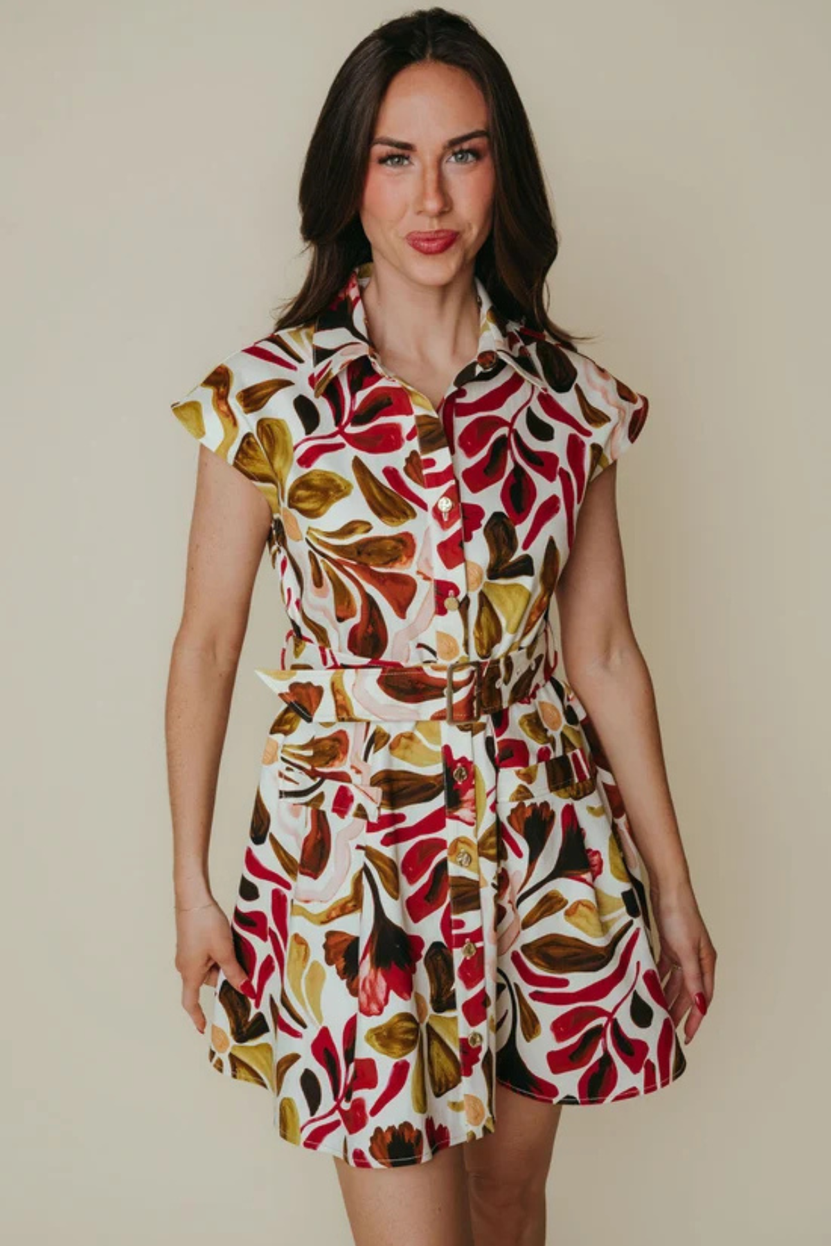 A woman modeling a short-sleeve collared dress with a bold retro leaf pattern in red, brown, olive green, and cream on a light background, set against a plain beige studio backdrop.