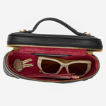 A black leather cosmetic case with a top handle, shown open to reveal a burgundy velvet interior. Inside, a pair of cream cat-eye sunglasses, a lip gloss tube, and a small gold keychain are neatly arranged.