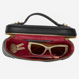 A black leather cosmetic case with a top handle, shown open to reveal a burgundy velvet interior. Inside, a pair of cream cat-eye sunglasses, a lip gloss tube, and a small gold keychain are neatly arranged.