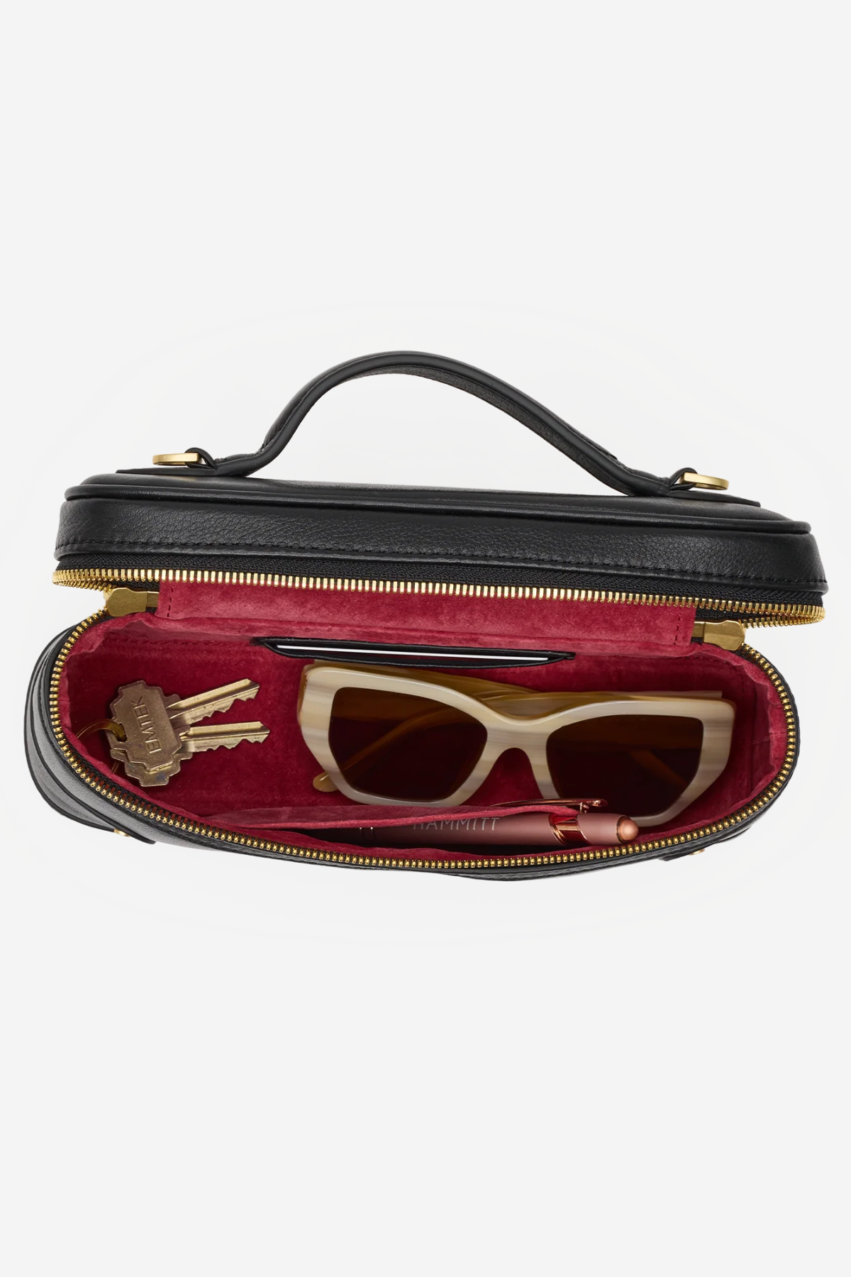 A black leather cosmetic case with a top handle, shown open to reveal a burgundy velvet interior. Inside, a pair of cream cat-eye sunglasses, a lip gloss tube, and a small gold keychain are neatly arranged.