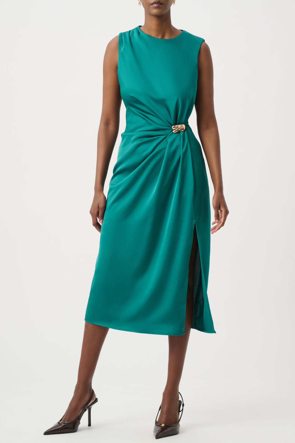 A woman models a sleeveless teal green midi dress with a twisted waist detail and a small gold-tone belt buckle, paired with dark brown pointed-toe heels against a light gray studio background.