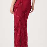 A woman wearing a floor-length red evening gown with floral lace applique and a front slit, shown from the back against a light gray studio background.