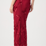A woman wearing a floor-length red evening gown with floral lace applique and a front slit, shown from the back against a light gray studio background.