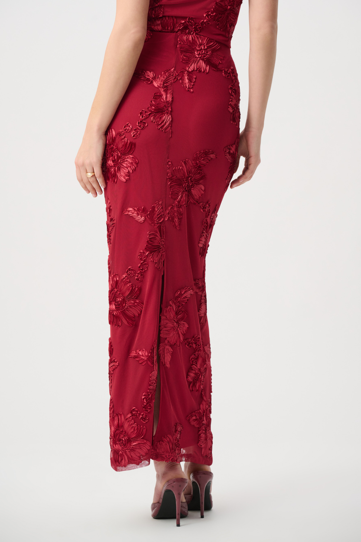 A woman wearing a floor-length red evening gown with floral lace applique and a front slit, shown from the back against a light gray studio background.