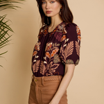 A woman modeling a short-sleeve blouse with a dark purple, orange, and brown floral pattern, paired with brown pants, set against a warm beige background with a green plant on the left.