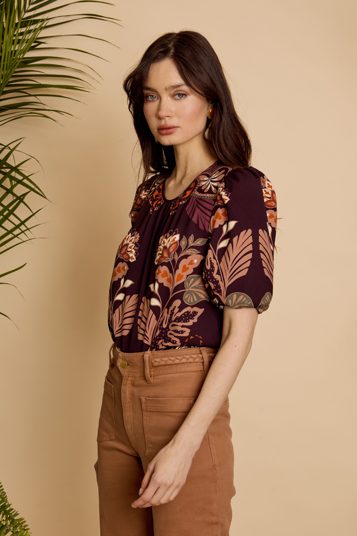 A woman modeling a short-sleeve blouse with a dark purple, orange, and brown floral pattern, paired with brown pants, set against a warm beige background with a green plant on the left.