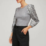 Woman wearing a gray top with leopard print sleeves and black pants on a plain background