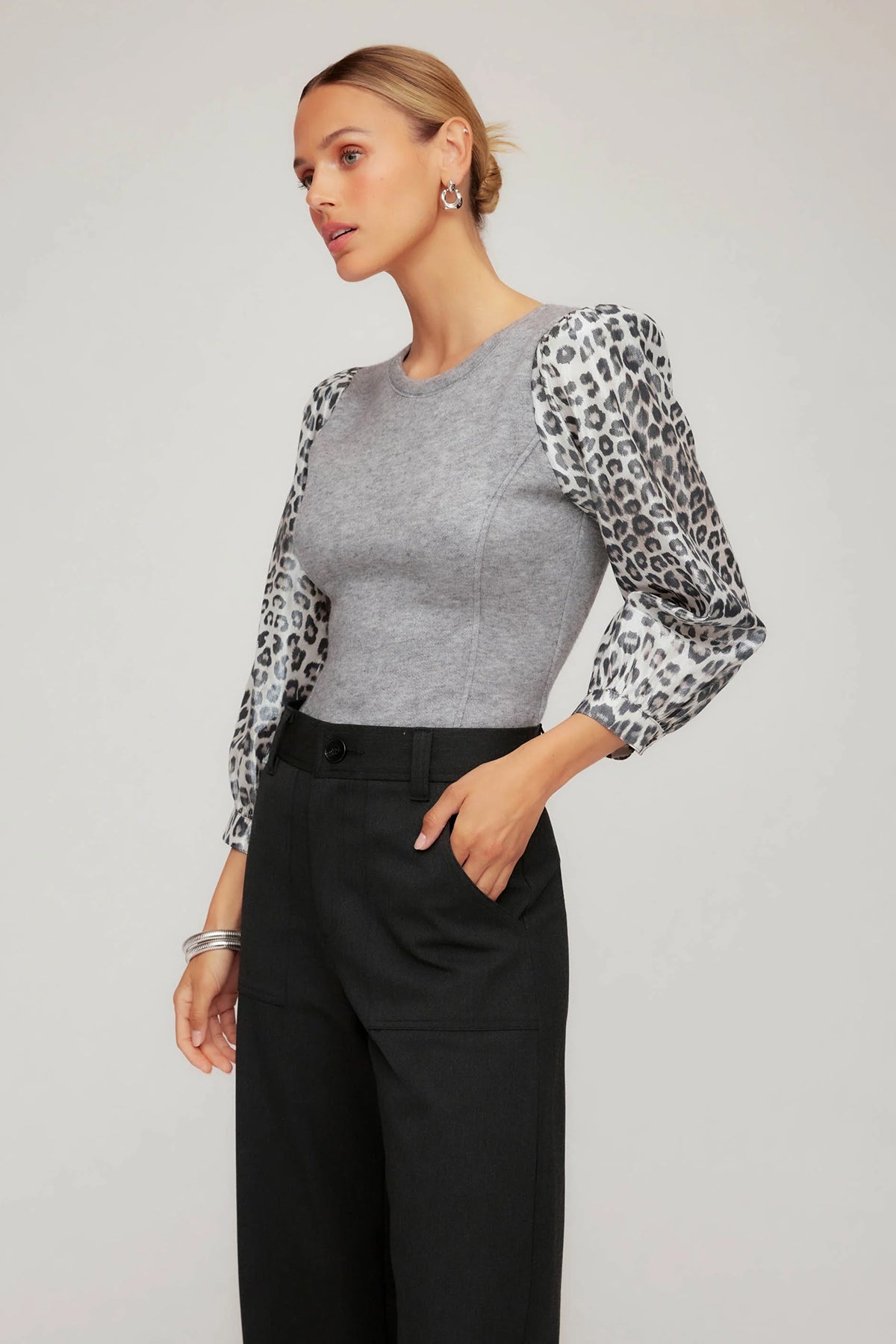 Woman wearing a gray top with leopard print sleeves and black pants on a plain background