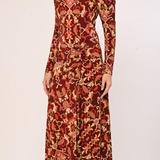 A woman wearing a long-sleeve midi dress with a dense abstract floral and paisley pattern in rust red, orange, brown, and beige, photographed against a plain light background.