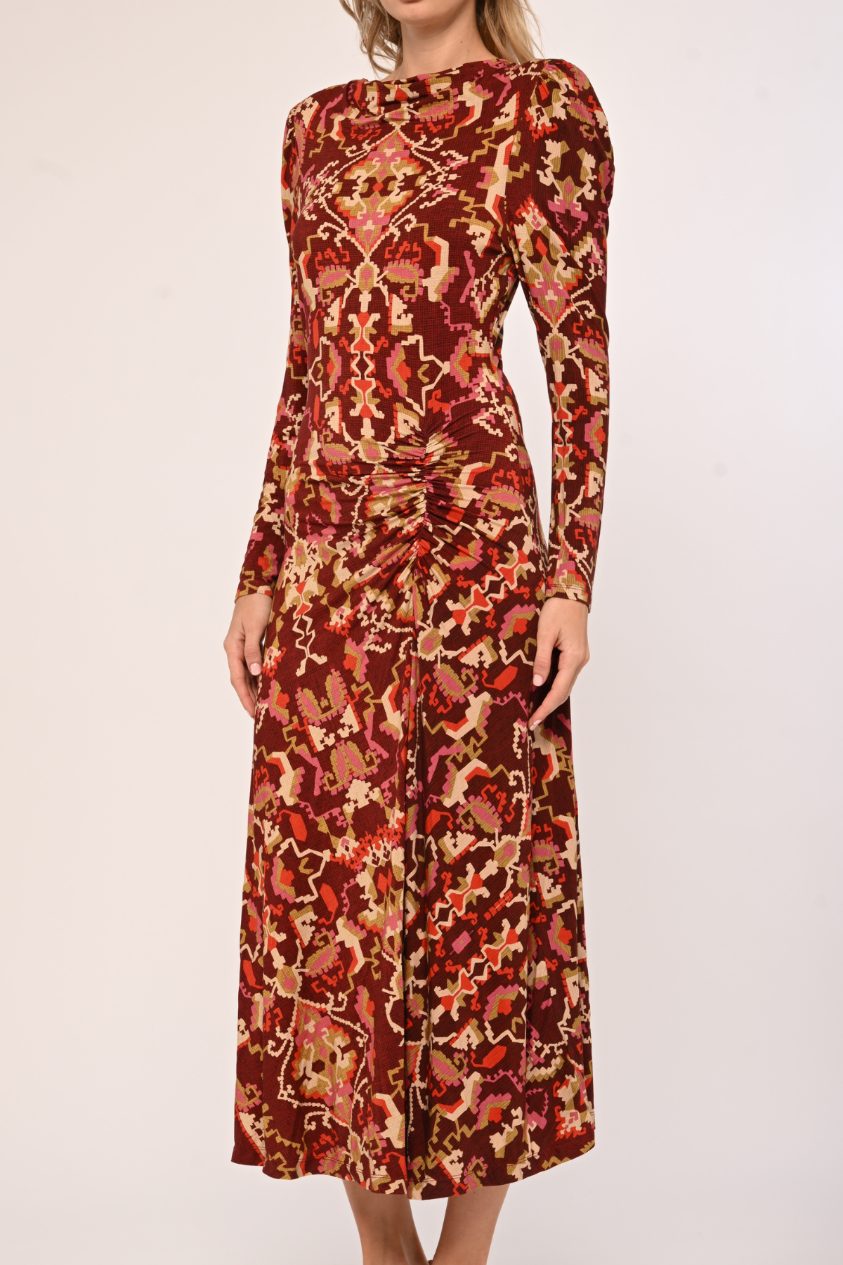 A woman wearing a long-sleeve midi dress with a dense abstract floral and paisley pattern in rust red, orange, brown, and beige, photographed against a plain light background.