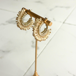A pair of gold crescent-shaped hoop earrings adorned with pearl-like beads, displayed on a gold stand against a white marble background.