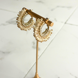 A pair of gold crescent-shaped hoop earrings adorned with pearl-like beads, displayed on a gold stand against a white marble background.