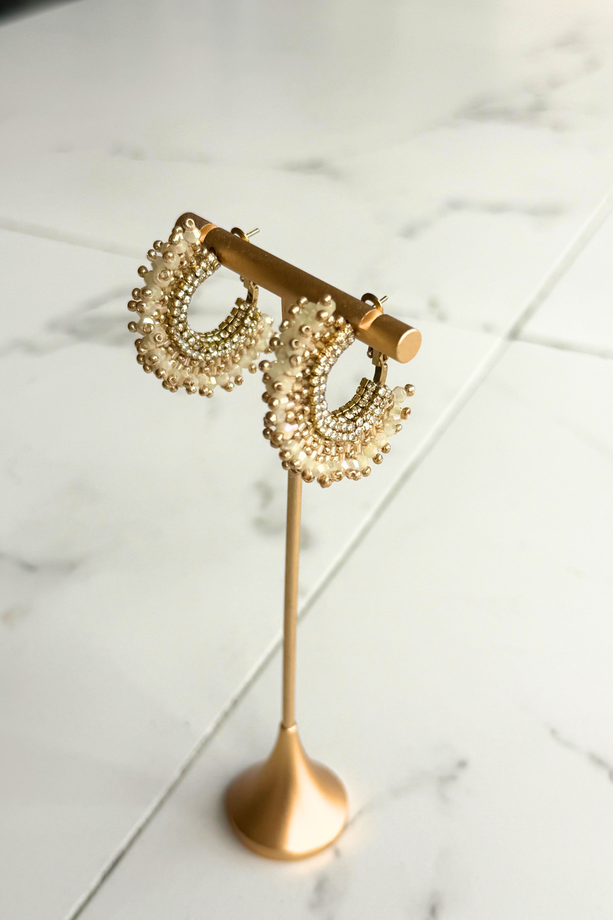 A pair of gold crescent-shaped hoop earrings adorned with pearl-like beads, displayed on a gold stand against a white marble background.