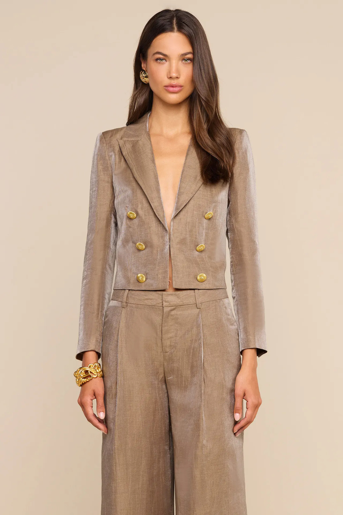 A woman modeling a taupe double-breasted blazer and matching wide-leg trousers with prominent gold buttons, set against a solid beige background.