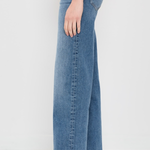 A side view of a person wearing medium-wash blue wide-leg jeans and beige woven slip-on shoes against a plain white studio background.