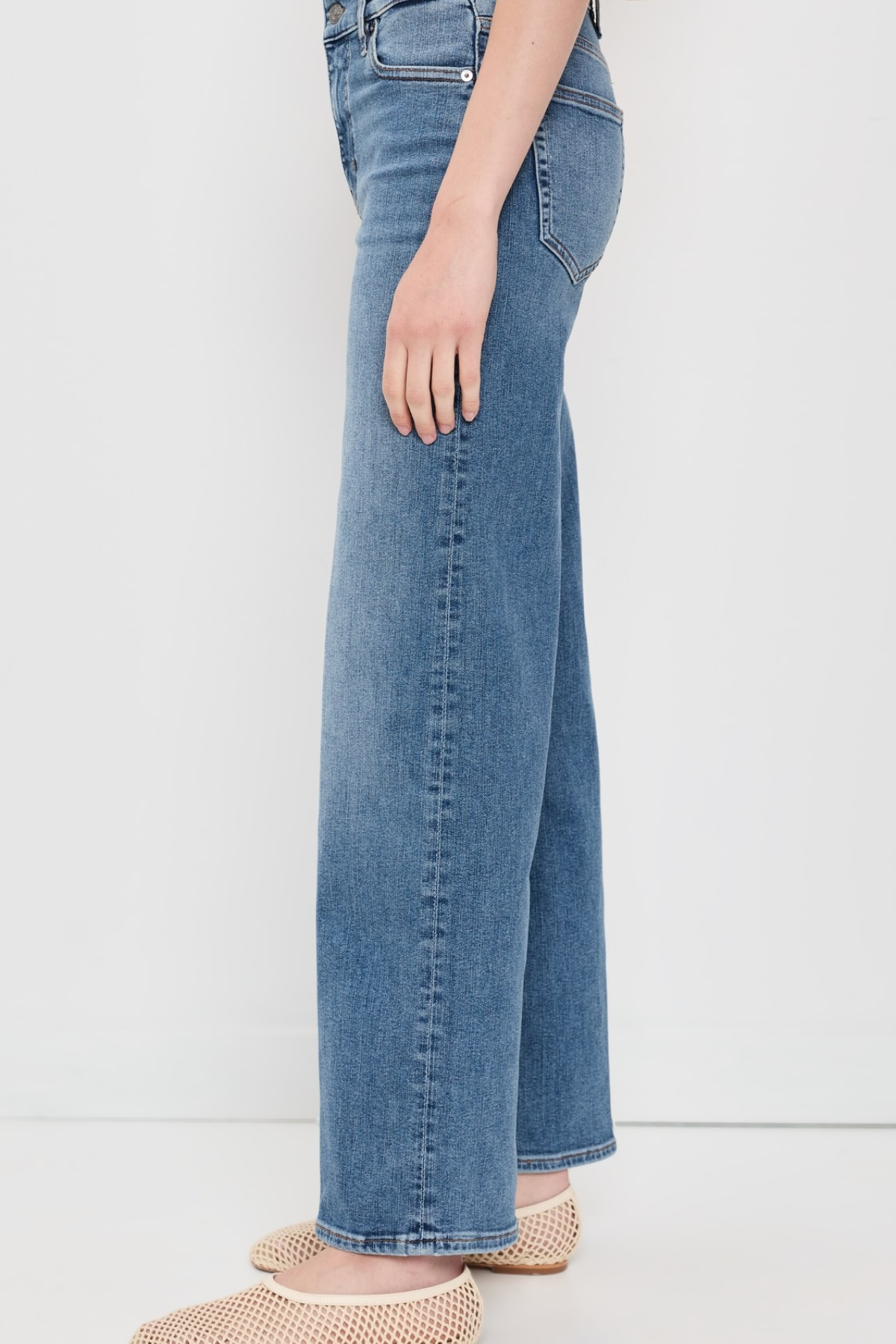 A side view of a person wearing medium-wash blue wide-leg jeans and beige woven slip-on shoes against a plain white studio background.