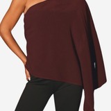 A woman wearing a dark burgundy one-shoulder knit poncho top with black leggings, shown from the neck down against a light gray background.