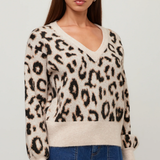 A woman wearing a beige and black leopard print knit sweater with a V-neck and blue jeans, standing against a light gray studio background.