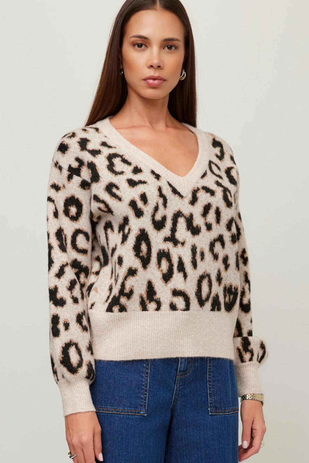 A woman wearing a beige and black leopard print knit sweater with a V-neck and blue jeans, standing against a light gray studio background.