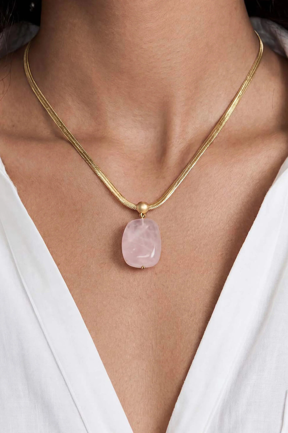 A close-up of a model wearing a delicate gold snake chain necklace with a smooth, rounded light pink stone pendant.