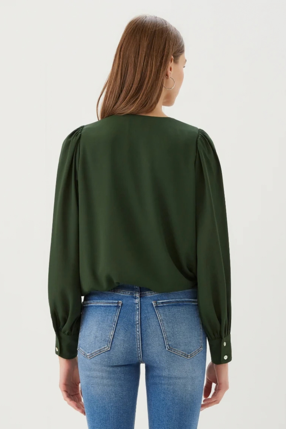 A woman viewed from the back wearing a dark green long-sleeve blouse with puffed sleeves and blue jeans, against a plain light gray studio background.