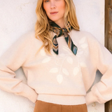 A woman wearing a cream knit sweater with a leaf pattern, a patterned neck scarf, and a brown skirt, posing against a textured white wall.