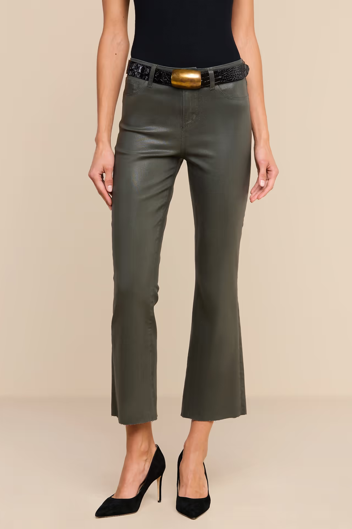 A model wearing dark olive green cropped flare pants with a black belt featuring a gold buckle, paired with a black top and black high heels, against a solid beige background.