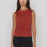 A woman modeling a sleeveless draped top in a muted terracotta or rust red color, paired with black pants, against a plain light gray studio background.