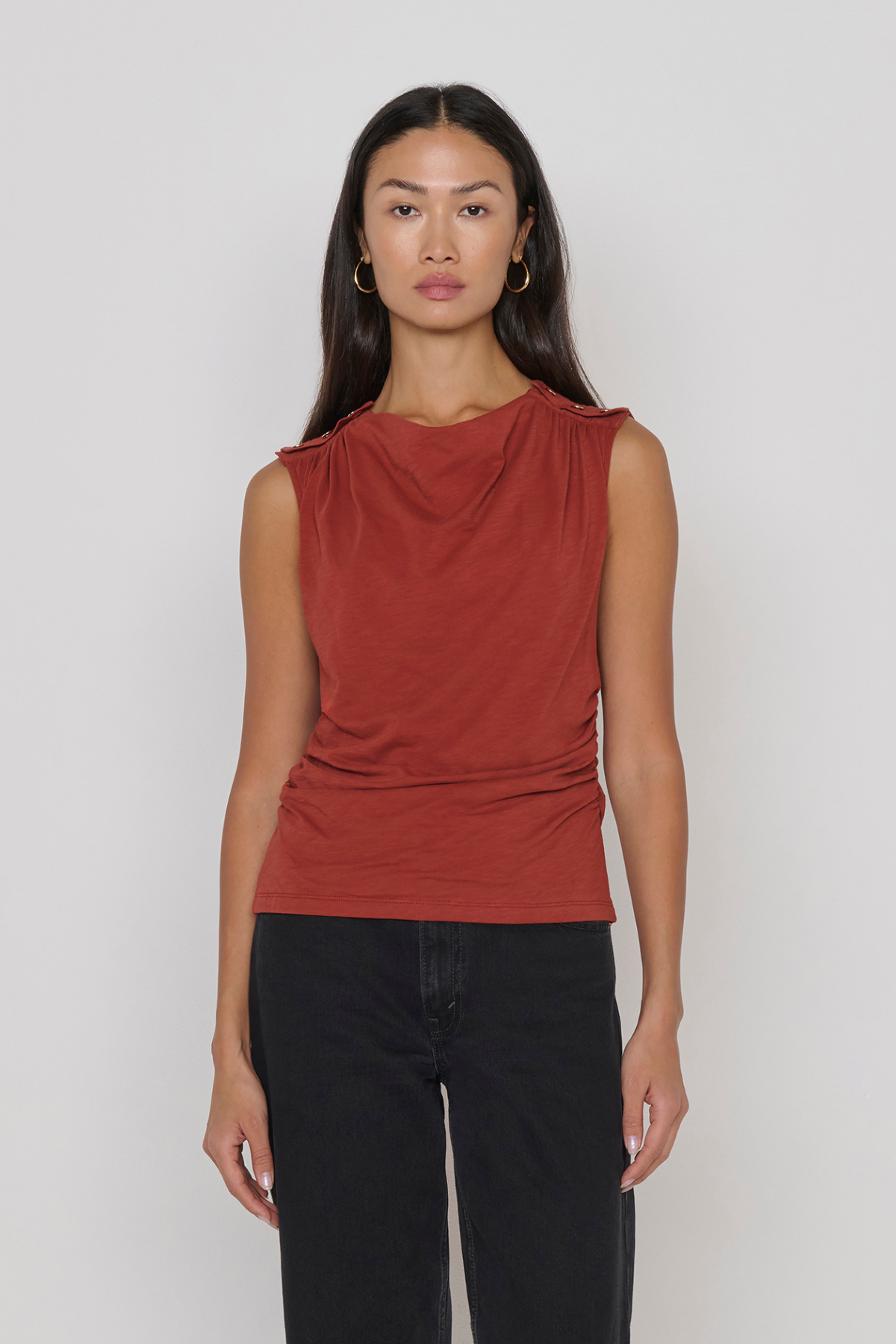 A woman modeling a sleeveless draped top in a muted terracotta or rust red color, paired with black pants, against a plain light gray studio background.