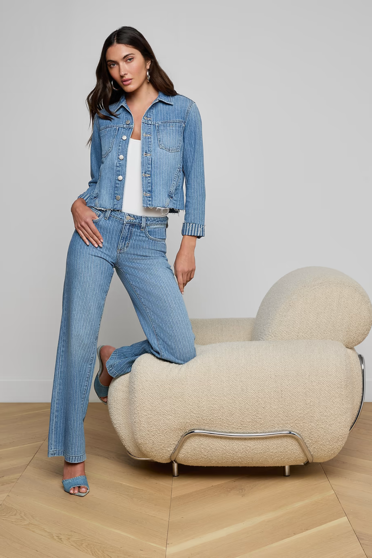 Woman in denim outfit standing next to a modern chair against a plain background