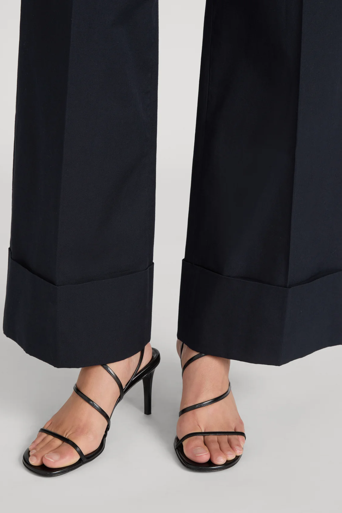 Black strappy high-heel sandals worn with dark navy wide-leg trousers, shown from the calves down on a light gray studio background.
