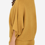A woman wearing a mustard yellow knit set featuring a draped, wide-sleeve top and matching pants, shown from a side-back angle against a plain light background.