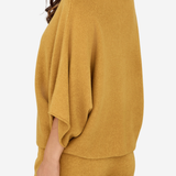 A woman wearing a mustard yellow knit set featuring a draped, wide-sleeve top and matching pants, shown from a side-back angle against a plain light background.