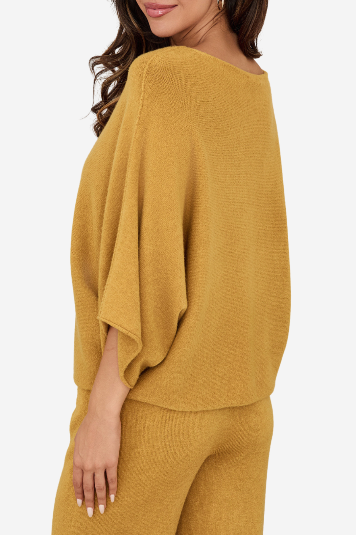 A woman wearing a mustard yellow knit set featuring a draped, wide-sleeve top and matching pants, shown from a side-back angle against a plain light background.