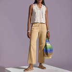 Woman in striped sleeveless top and beige pants holding a colorful bag on a gray platform with purple background