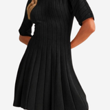 A woman wearing a black pleated short-sleeve dress with a crew neck, shown from the shoulders to mid-thigh against a plain light background.