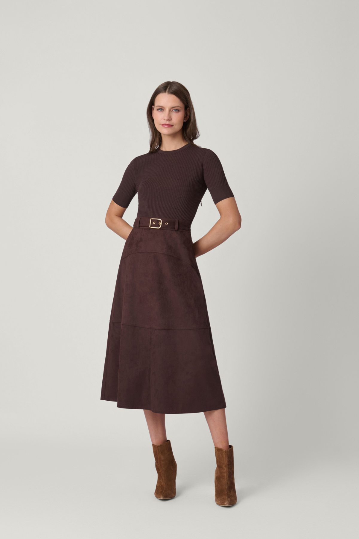 Woman wearing a brown dress with a belt and brown boots on a light gray background