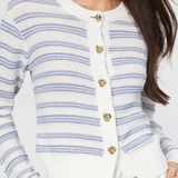 Woman wearing a white cardigan with blue stripes and gold buttons on a light gray background