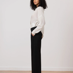 Woman wearing a white blouse and black pants standing against a plain white wall.