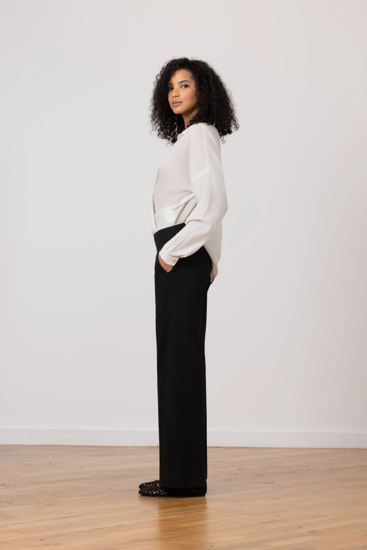 Woman wearing a white blouse and black pants standing against a plain white wall.
