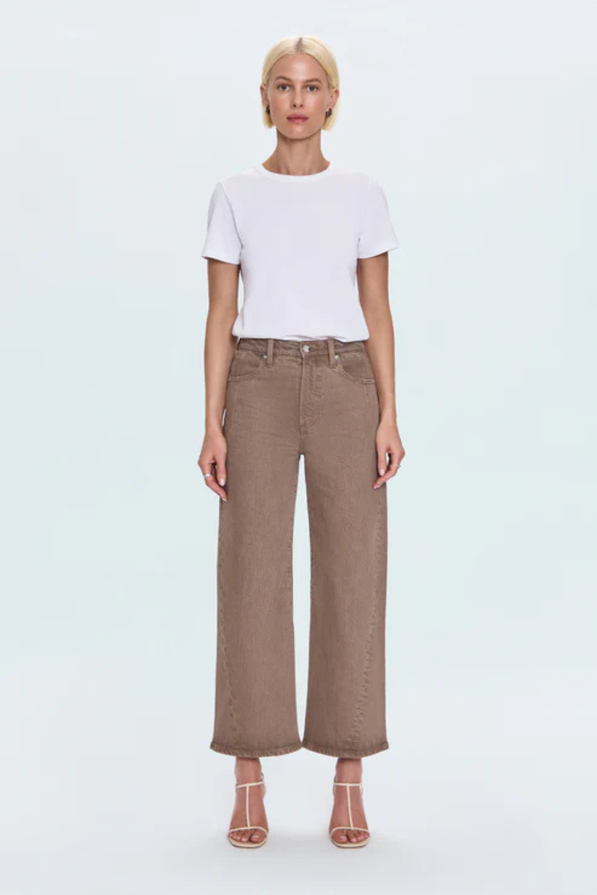 A model stands against a light gray studio background wearing a white short-sleeve crewneck t-shirt tucked into high-waisted, wide-leg brown cropped jeans, paired with beige strappy high-heeled sandals.